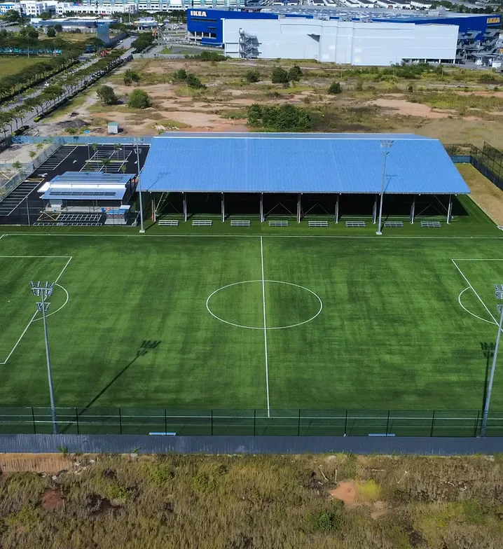 Aerial daylight view of Batu Kawan showing the blue-roofed covered pitch and the full-size 11-a-side pitch, with the IKEA Batu Kawan store behind