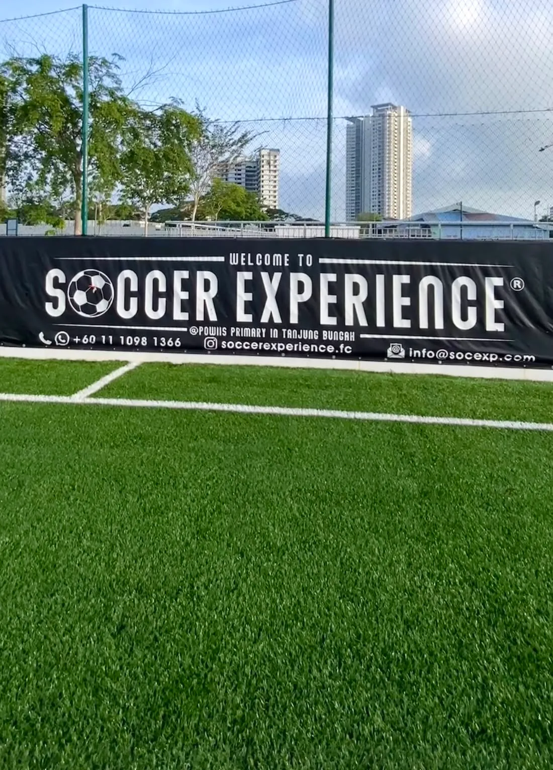 The Welcome to Soccer Experience banner along the perimeter fence at Tanjung Bungah, with the turf in the foreground and the towers of Tanjung Tokong behind