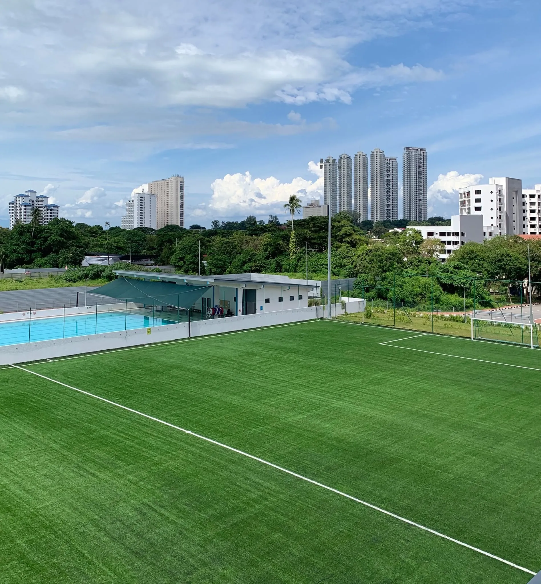 The 9-a-side pitch at Soccer Experience Tanjung Bungah in daylight, with the poolside clubhouse alongside it and the towers of Tanjung Tokong behind
